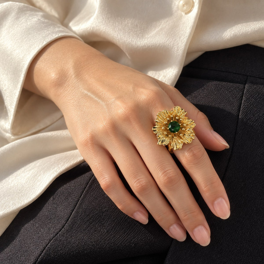 Gold-tone flower ring with a green rhinestone worn on a hand with a cream blouse and dark trousers.